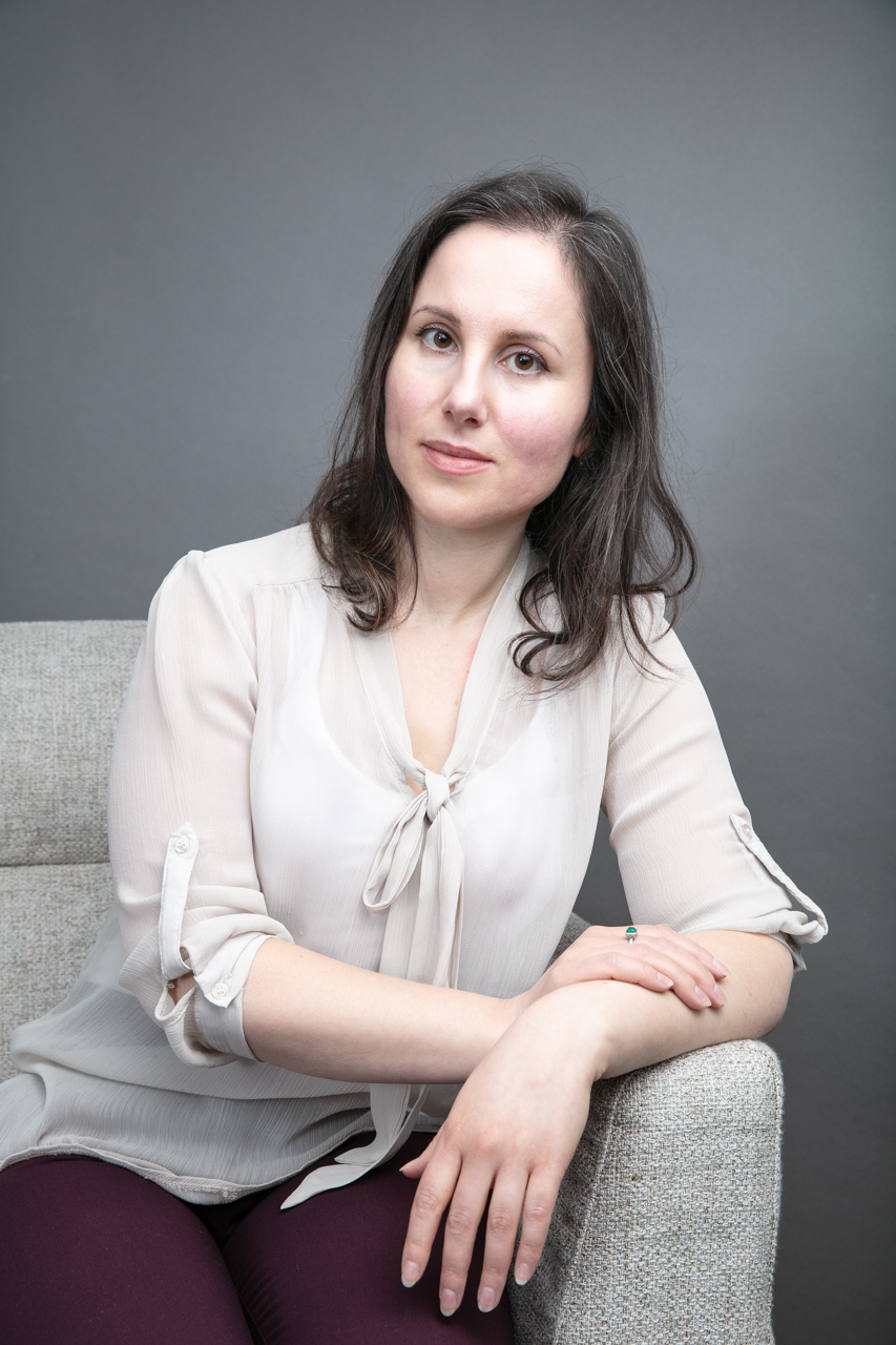 Portrait of a woman with dark hair in a light blouse seated on a gray textured chair against a neutral backdrop, looking at the camera.